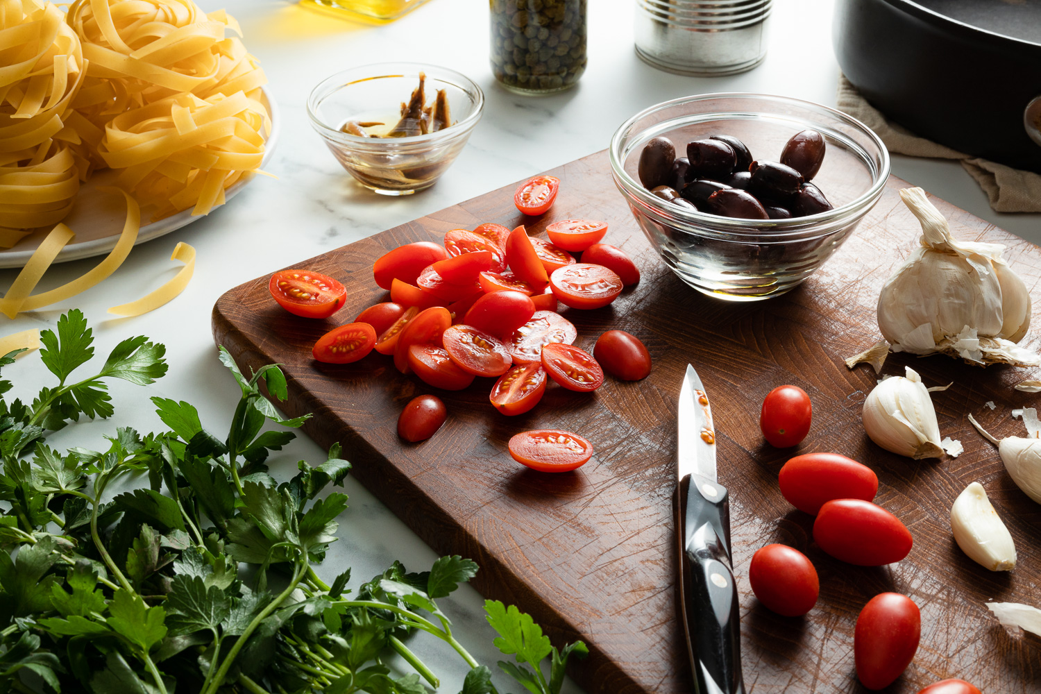 tomatoes, olives, garlic, and a paring knife on a cutting board with parsley and pasta